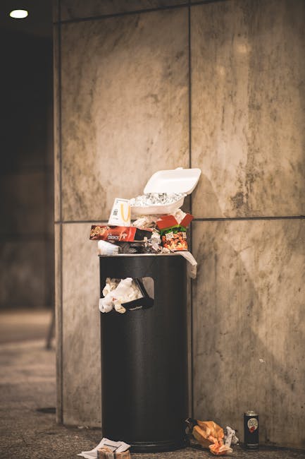A person's arm extends from the left side of the image, holding a green and blue plastic bag loosely tied at the top, which is filled with assorted waste materials. The bag appears slightly translucen