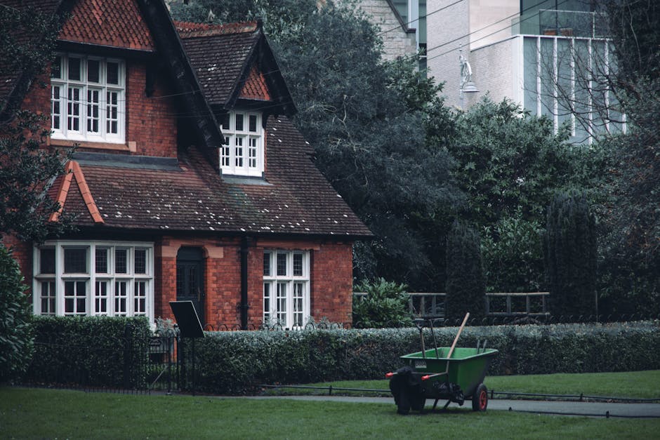 A green wheelbarrow with red handles and wheels is positioned on a paved pathway in the front garden of a residential property. The wheelbarrow is filled with long-handled gardening tools, including a