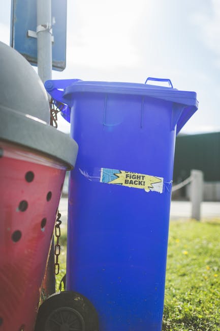 A sanitation worker dressed in a red high-visibility vest and black trousers is standing near the rear of a large red waste collection truck on a roadside. The worker appears to be handling black garb