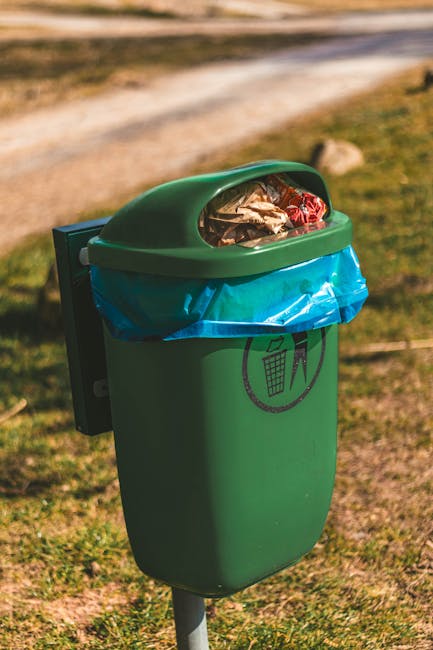 A row of large, wheeled wheelie bins positioned along a paved pathway next to a brick wall, with each bin featuring a distinct colour lid—green, red, blue, and dark green—indicating different type