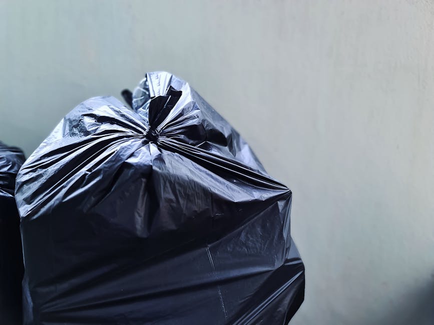 A large collection of overflowing rubbish bins and waste bags situated on a paved sidewalk in front of a commercial area, with two black wheelie bins and a red bin among them. Various items such as ca
